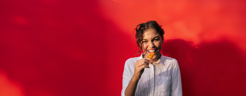 Junge Frau mit Locken und einem Stück Mango in der Hand lacht vor einem Roten Hintergrund in die Kamera